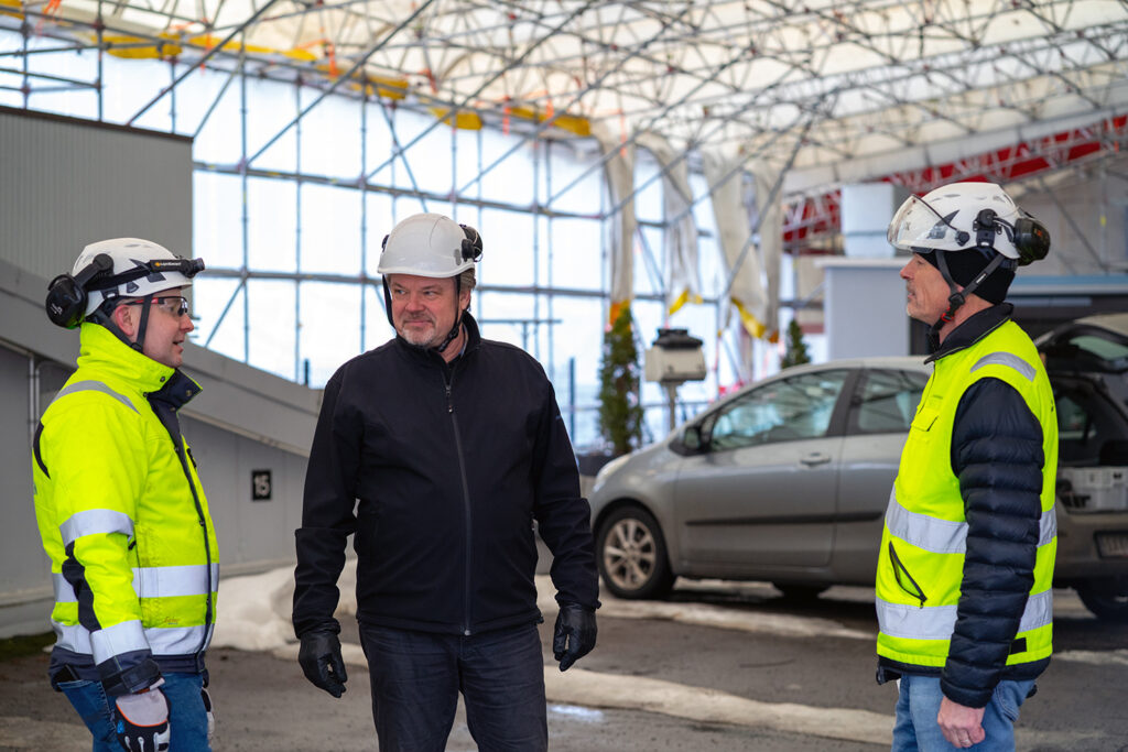 Kolme miestä työmaalla valkoisissa suojakypärissä ja suojavaatetuksessa keskustelemassa taloyhtiön sisäpihalla